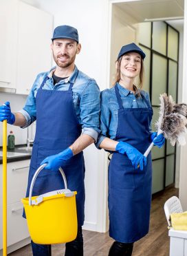Portrait of a couple as a professional cleaners in uniform standing together with cleaning tools indoors Portrait of a couple as a professional cleaners in uniform standing together with cleaning tools indoors